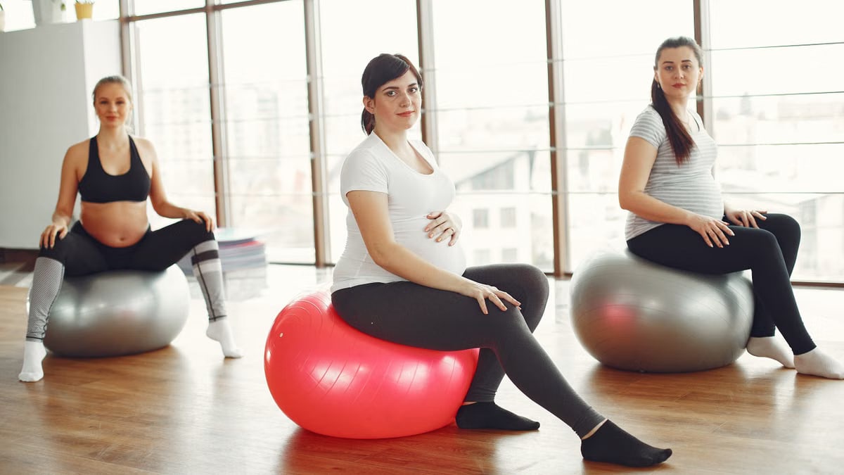 Positive pregnant ladies sitting on fit balls during prenatal training in spacious light gym at daytime and smiling while looking at camera