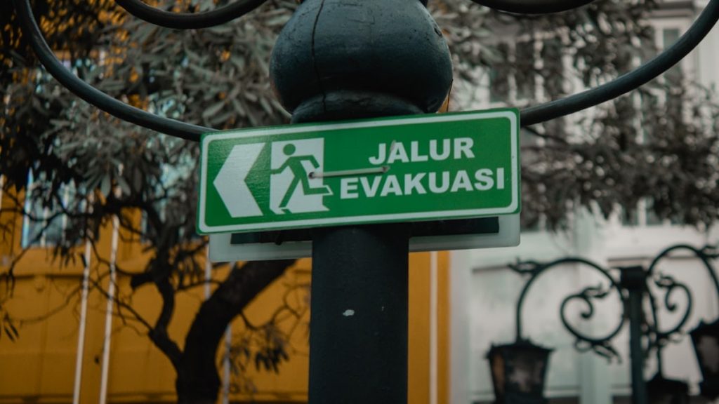 A bright green rectangular sign reading "JALUR EVAKUASI" (Evacuation Route) hangs from a weathered black metal pole adorned with intricate curved brackets. The sign features a bold white pictogram of a running figure, directing attention to the left. Set against a softly blurred backdrop of leafy branches and a yellow-walled building with ornate iron railings, the image balances functional urban infrastructure with subtle artistic detail, emphasizing clarity and safety in public spaces.