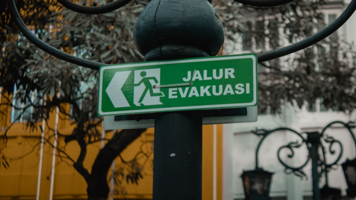 A bright green rectangular sign reading "JALUR EVAKUASI" (Evacuation Route) hangs from a weathered black metal pole adorned with intricate curved brackets. The sign features a bold white pictogram of a running figure, directing attention to the left. Set against a softly blurred backdrop of leafy branches and a yellow-walled building with ornate iron railings, the image balances functional urban infrastructure with subtle artistic detail, emphasizing clarity and safety in public spaces.